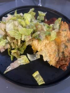 A black plate with a serving of leafy green salad and a portion of savory Dutch baby omelet. The salad appears lightly dressed, and the fluffy omelet has visible herbs mixed in.