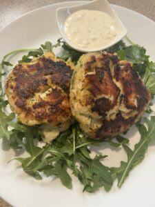 Two golden-brown fish cakes served on a bed of fresh arugula, accompanied by a small dish of creamy dipping sauce on a white plate.