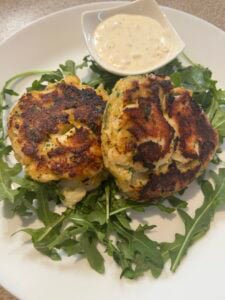 Two golden-brown fish cakes rest on a bed of fresh arugula, served with a small dish of creamy dipping sauce on a white plate.