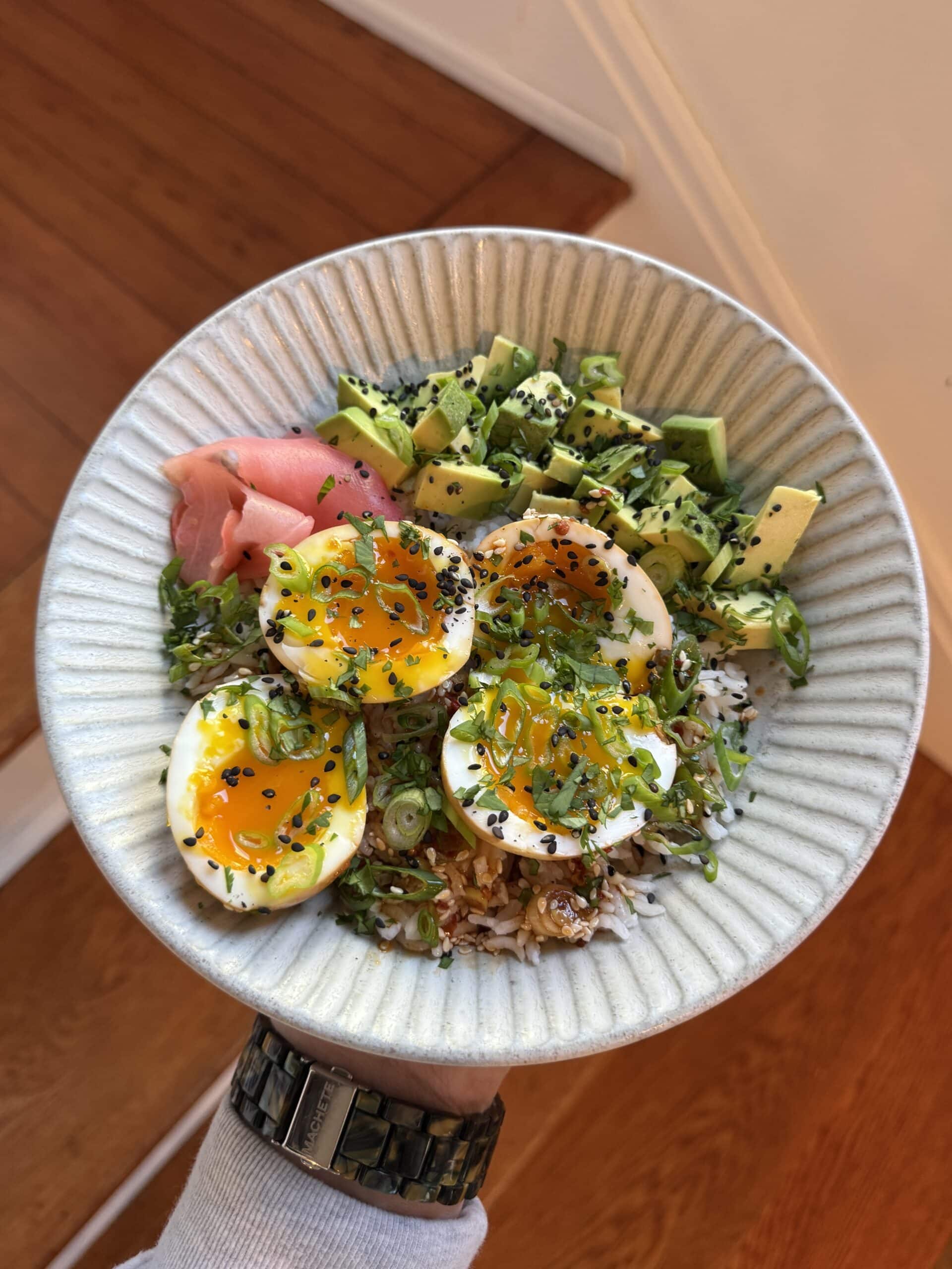 A hand holds a bowl of rice topped with sliced soy sauce eggs, diced avocado, pickled ginger, chopped scallions, black and white sesame seeds, and shredded nori. The background features a wooden floor and light-colored wall.