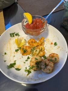 A white plate with old bay steamed shrimp garnished with chopped parsley and a lemon wedge, next to a glass dish of cocktail sauce with a spoon and a lemon slice on the rim.