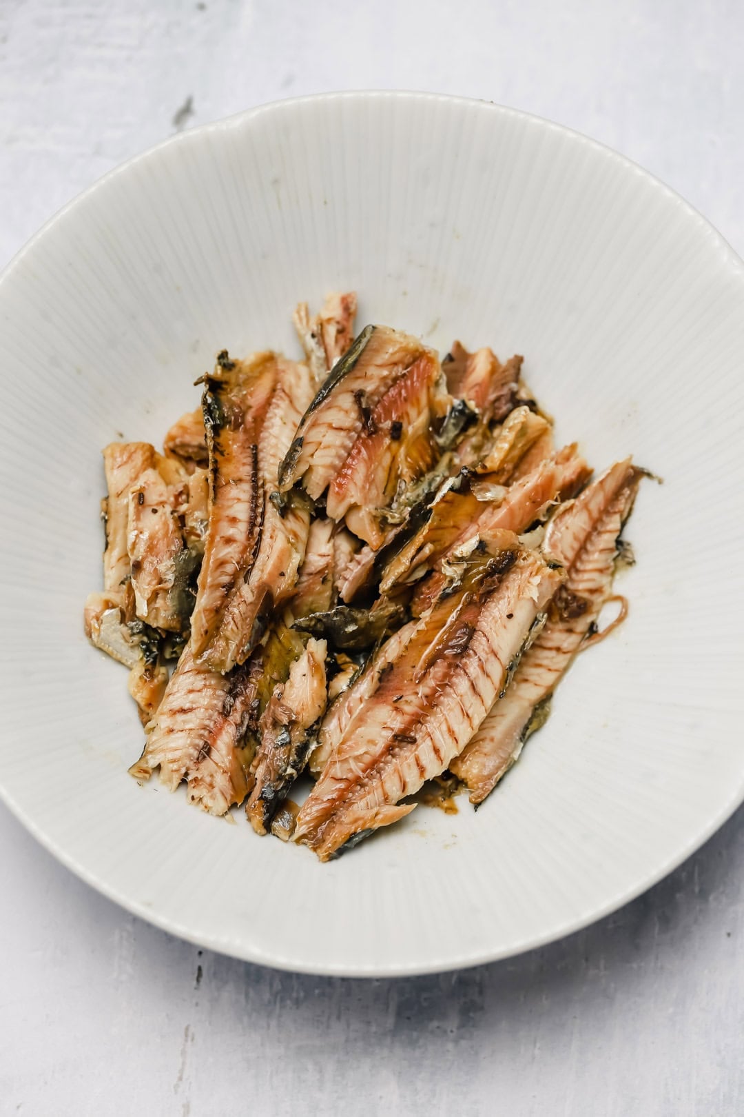 Shredded cooked fish pieces with skin and bones are arranged in a white, shallow bowl on a light surface, reminiscent of rustic sardine rillettes.