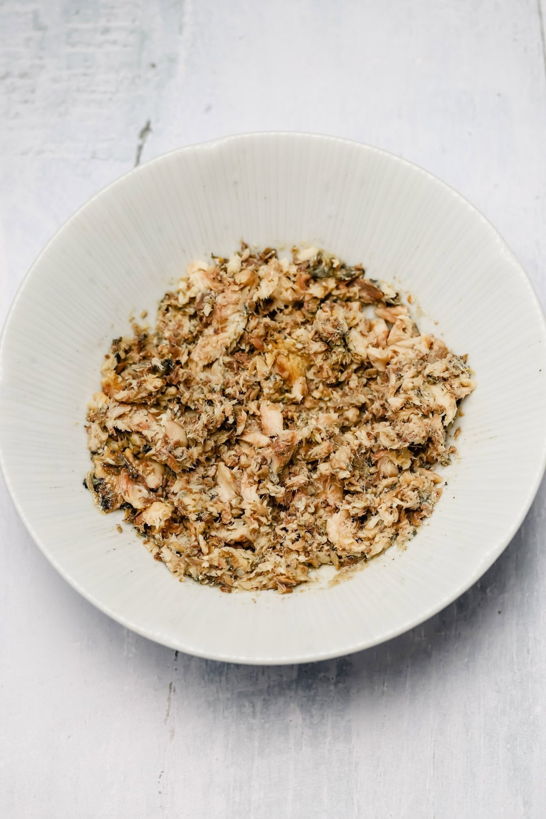 A white bowl filled with flaked, seasoned fish sits on a light, textured surface. The sardine rillettes are mixed with herbs and appear finely shredded.