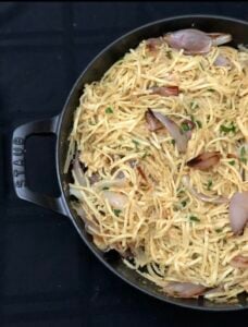 A black Staub pan filled with caramelized shallot pasta and chopped parsley, set on a dark background.
