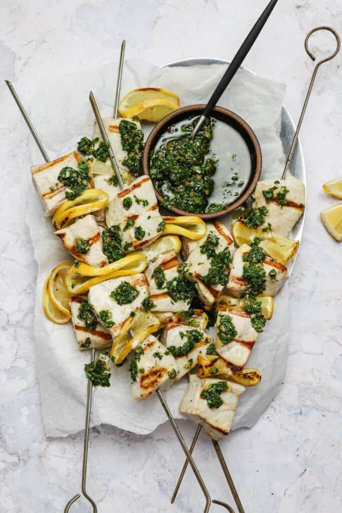Grilled swordfish skewers on a tray with Italian salsa verde spooned on top.