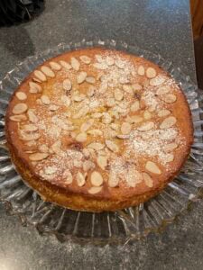 A round almond ricotta cake sits on a glass plate, topped with sliced almonds and a dusting of powdered sugar, displayed on a dark countertop.