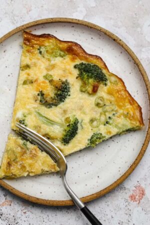 Slice of crustless broccoli cheddar quiche on a plate with a fork slicing it.