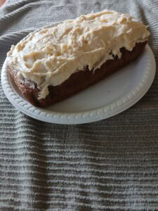 A rectangular carrot cake loaf with creamy white frosting sits on a white plastic plate, placed on a textured gray fabric background.