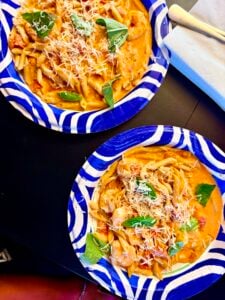 Two blue and white plates filled with creamy Penne Alla Vodka, topped with grated cheese and fresh basil leaves, are on a dark table beside a napkin and silverware.