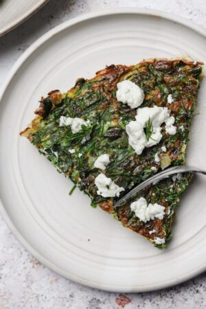 Cutting into a slice of spinach frittata with goat cheese on a white plate.