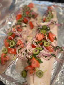 Two raw Mediterranean baked cod fillets on a foil-lined baking sheet topped with chopped tomatoes, sliced green olives, red onion, and herbs.