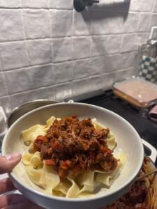 A hand holds a bowl of wide pasta noodles topped with chunky braised lamb ragu in a kitchen with a tiled backsplash and cooking utensils in the background.