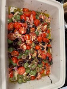 A baking dish filled with roasted cherry tomatoes, green and black olives, sliced red onions, garlic, and tender mediterranean baked cod, all mixed in olive oil.