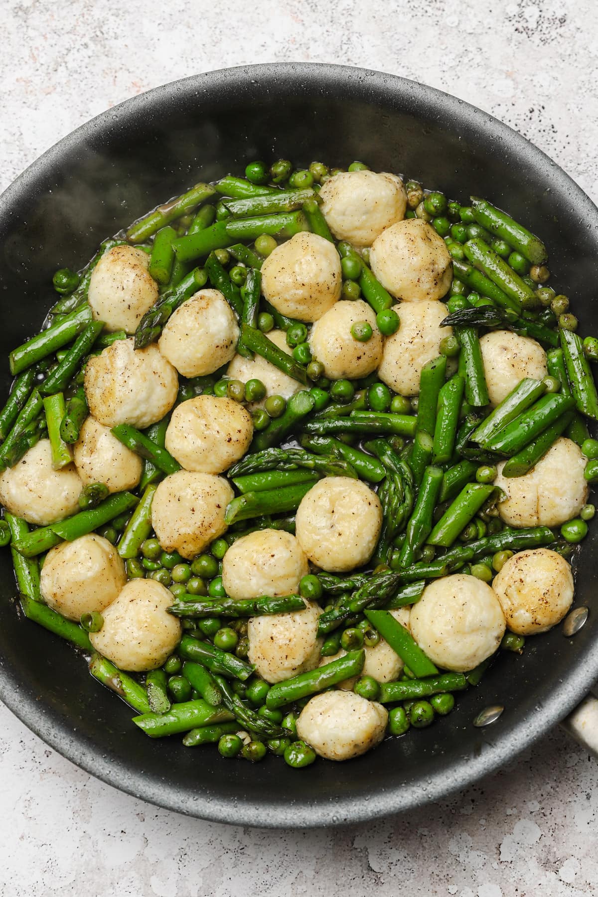 Ricotta gnudi recipe cooking in a skillet with browned butter and asparagus and peas.