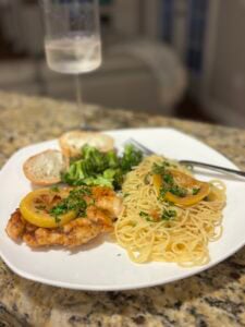 A white plate with chicken piccata, pasta garnished with parsley, steamed broccoli, and a slice of bread; a fork and a glass of white wine rest in the background on a granite countertop.