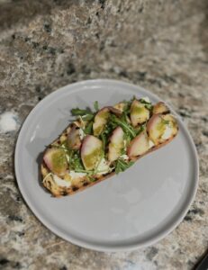 A slice of toasted bread topped with cheese, arugula, sliced radishes, and a vibrant basil vinaigrette is served on a gray plate atop a granite countertop.