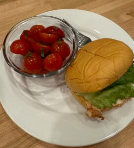 A white plate with a chicken sandwich on a bun with lettuce sits beside a glass bowl of marinated tomatoes mixed with herbs and a fork. The plate is on a wooden surface.