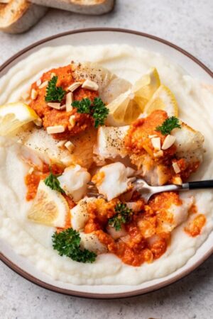 Fork cutting pan seared cod with romesco sauce on bed of mashed potatoes.