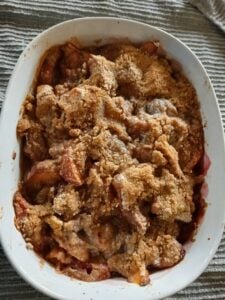 A baked peach crumble in a white dish, topped with a golden-brown, crumbly layer. The fruit filling is bubbling around the edges. The dish is placed on a striped fabric surface.