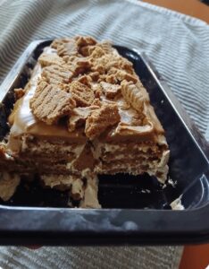 A rectangular cookie butter icebox cake with layers of cream and cookies, topped with crumbled cookies, sits in a black tray with a portion missing. The tray rests on a striped fabric surface.