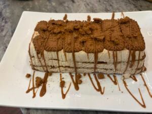 A rectangular layered dessert with cookies and cream, inspired by a classic cookie butter icebox cake, topped with crushed cookies and a drizzle of caramel sauce, served on a white rectangular plate.