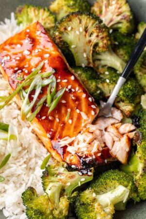 Fork cutting teriyaki salmon on bed of rice with side of broccoli.