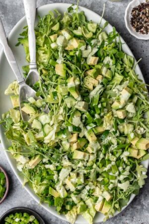 Overhead view of serving spoons holding green goddess salad tossed in dressing on large plate.