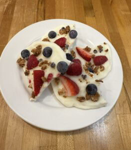 A white plate holds three pieces of greek yogurt bark topped with strawberries, blueberries, raspberries, and granola, set on a wooden table.