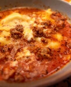 A close-up of lasagna soup with creamy melted cheese, ground meat, and rich red tomato sauce in a shallow bowl. The surface appears seasoned with herbs and spices, evoking the comforting flavors of classic baked lasagna.