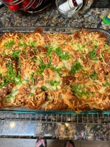 A glass baking dish filled with a golden-brown croissant breakfast casserole, topped with melted cheese and chopped green herbs, rests on a cooling rack on a colorful kitchen countertop.