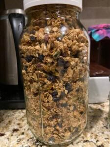 A glass jar filled with granola, nuts, seeds, and dried fruit sits on a kitchen counter. The jar has a white lid, and kitchen appliances and a colorful towel are visible in the background—reminding one of the delightful flavors of Eleven Madison Park’s granola.