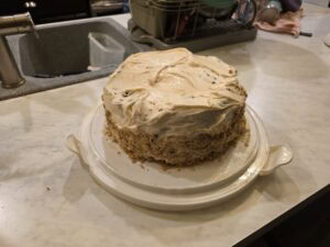 A homemade round carrot cake with cream cheese frosting sits on a white cake stand on a kitchen counter, with a sink and dish rack visible in the background.