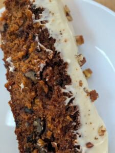 A close-up of a slice of carrot cake with cream cheese frosting, visible walnuts, and small bits of chopped nuts, served on a white plate.