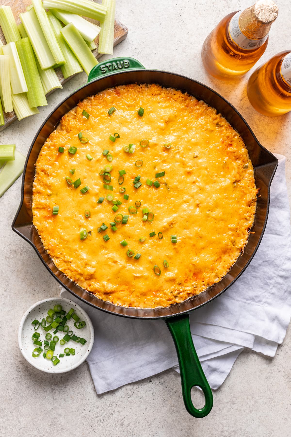 Baked buffalo chicken dip in a cast-iron skillet with thinly sliced scallions sprinkled on top, surrounded by a couple of beers, celery sticks,  and white linen napkin.