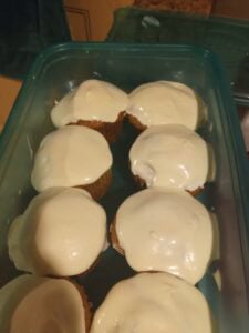 Eight cupcakes in a blue plastic container, each topped with a thick layer of smooth white icing reminiscent of carrot cake with cream cheese frosting. The container rests on a countertop.