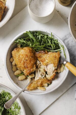Roasted chicken thighs in a bowl with butter beans and arugula salad.