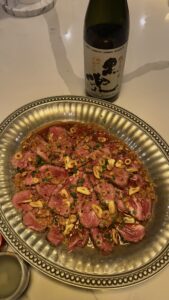 A round metal plate of beef tataki, filled with thinly sliced seared beef topped with chopped green onions and garlic chips, is served with sauce on a marble counter next to a bottle of sake.