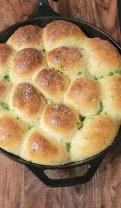 Golden, fluffy pull-apart rolls baked in a black cast-iron skillet, sprinkled with grated cheese and chopped parsley, rest invitingly on a wooden surface.