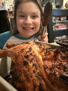 A smiling child holds tongs behind a large, roast tarragon chicken—beautifully roasted and herb-seasoned—in a welcoming kitchen setting.