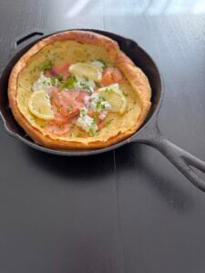 A smoked salmon Dutch baby in a cast iron skillet, topped with lemon slices, sour cream, and fresh chives, rests on a dark wooden table bathed in natural light.