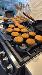 Two muffin trays filled with freshly baked carrot cake cupcakes rest on a stovetop, beside a black frying pan and various kitchen items. The kitchen scene looks bright and inviting.