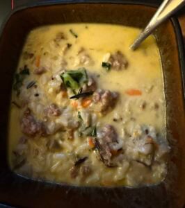A bowl of creamy soup with chunks of chicken sausage and wild rice, chopped greens, and visible pieces of carrot, with a spoon resting inside.