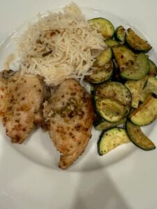 A plate with two pieces of creamy dijon chicken, a serving of white rice, and sautéed zucchini slices, all arranged separately on a white plate.