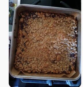 A square metal baking pan filled with a mixed berry fruit crisp, featuring a golden-brown, crumbly topping, sits on a stovetop. The crisp reveals a vibrant fruit filling beneath the topping.