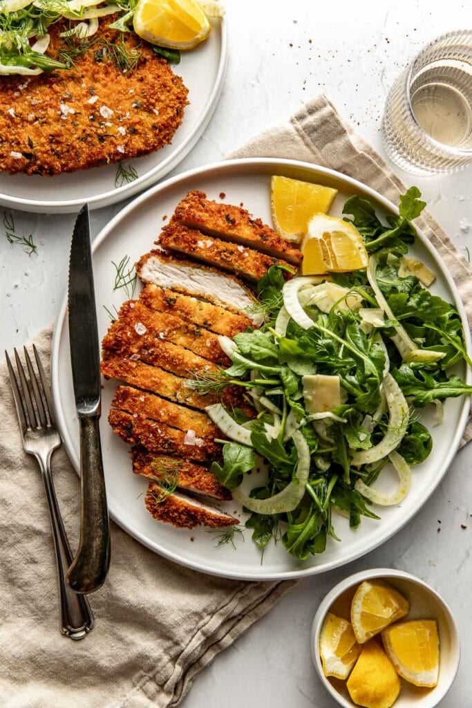 A white plate with sliced breaded chicken cutlet, fresh arugula and fennel salad, avocado, and lemon wedges. A glass of water, fork and knife, napkin, and a bowl with extra lemon wedges are nearby on a light surface.