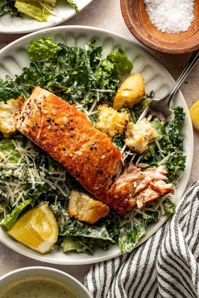 A plate of Caesar salad topped with a grilled salmon fillet, shredded parmesan cheese, croutons, and a lemon wedge. A fork holds a piece of crouton. A striped napkin and salt bowl are nearby.