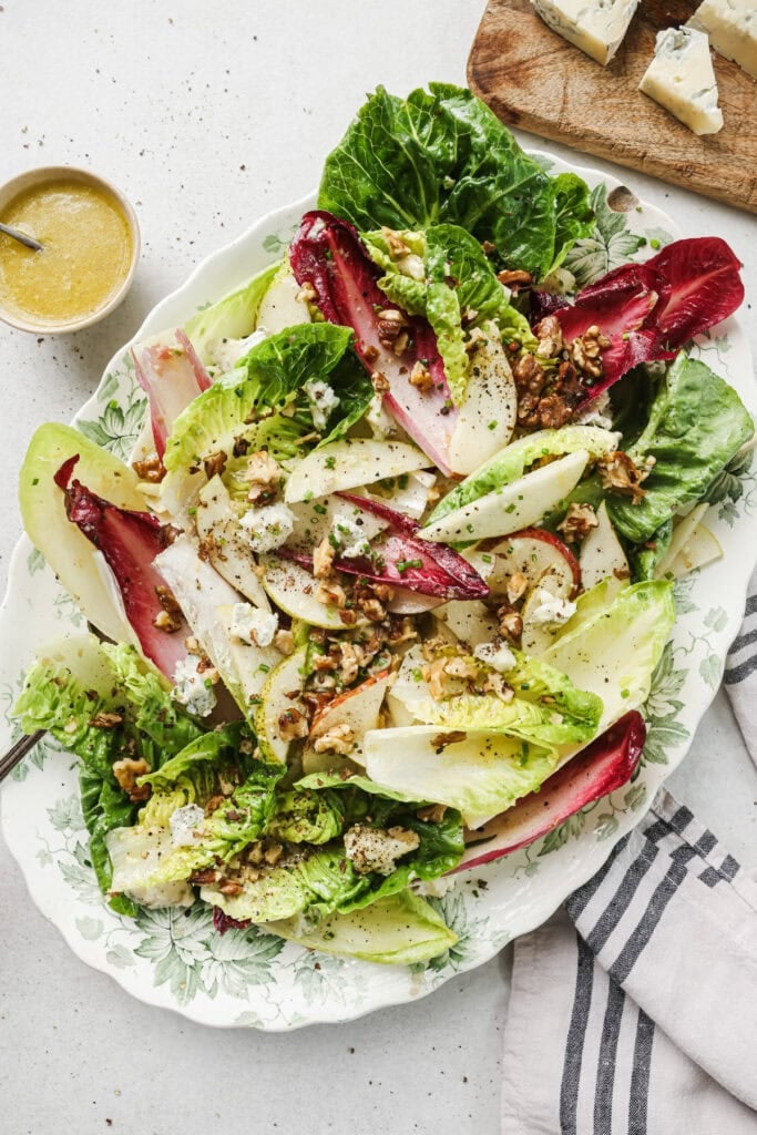 A platter of mixed green and red leafy salad with crumbled blue cheese, walnuts, and cracked black pepper, next to a small bowl of dressing and a wedge of blue cheese on a wooden board. A striped napkin is nearby.