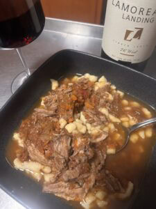 A black bowl filled with shredded beef in a rich tomato-based sauce over small dumplings, reminiscent of duck leg ragu, with a spoon inside. A glass of red wine and a bottle labeled Lamoreaux Landing sit beside the bowl.
