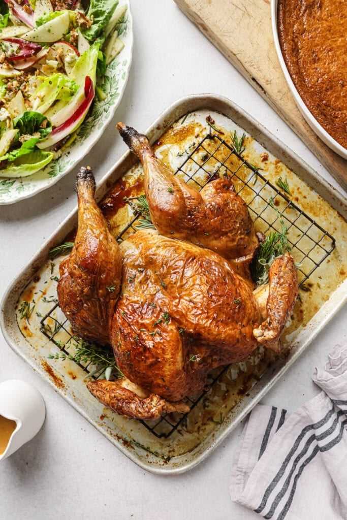 A golden-brown roasted chicken rests on a rack in a baking tray with herbs underneath. Nearby are a plate of salad, a dish of cornbread, a pitcher of gravy, and a striped towel.