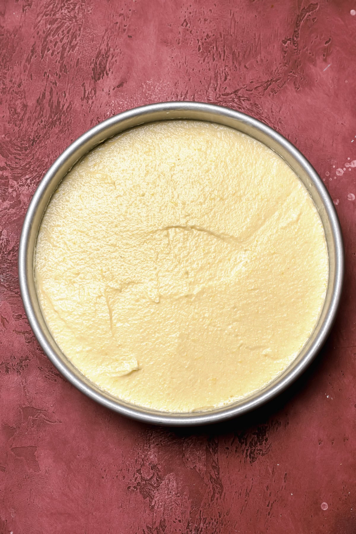 A round cake pan filled with smooth, uncooked yellow batter sits on a textured reddish-brown surface.